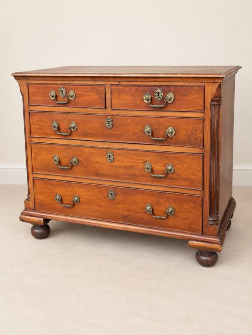 fig 1. A small Georgian mahogany chest of drawers featuring two short over three long drawers with decorative brass handles, fluted quarter columns, and turned bun feet.