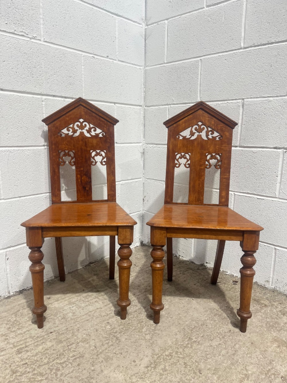 A pair of 19th-century carved oak hall chairs with pointed pediment tops, pierced fretwork back splats, flat timber seats, and turned front legs. View II