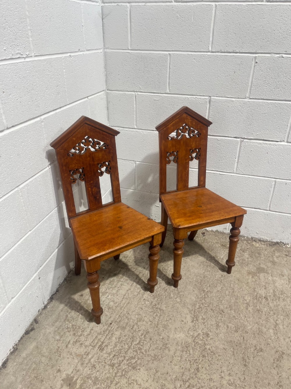 A pair of 19th-century carved oak hall chairs with pointed pediment tops, pierced fretwork back splats, flat timber seats, and turned front legs. View V