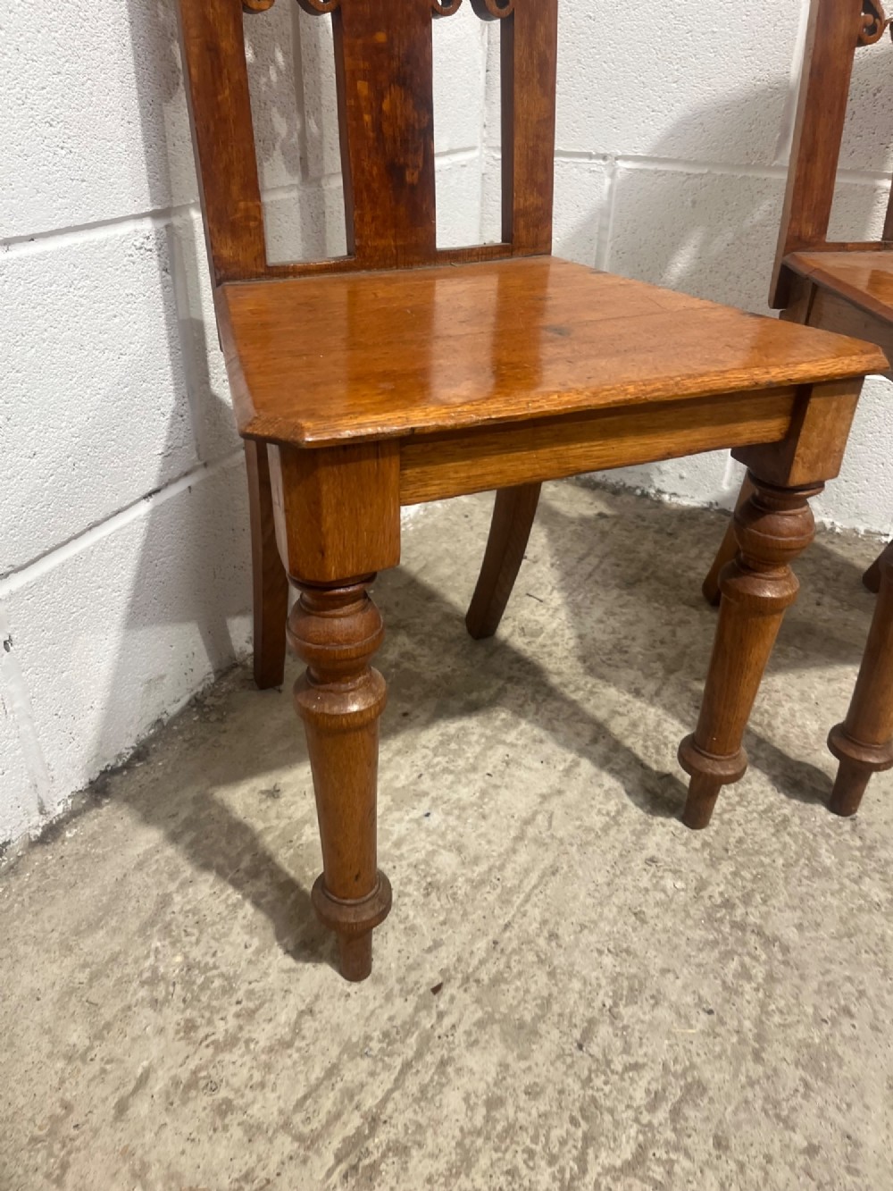 A pair of 19th-century carved oak hall chairs with pointed pediment tops, pierced fretwork back splats, flat timber seats, and turned front legs. View VII