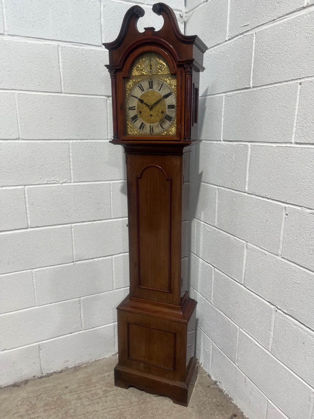 An antique Edwardian mahogany longcase clock featuring a swan-neck pediment, brass arch dial with Roman numerals, and an arched trunk door. View II