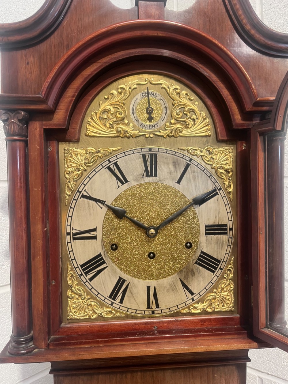 An antique Edwardian mahogany longcase clock featuring a swan-neck pediment, brass arch dial with Roman numerals, and an arched trunk door. View V