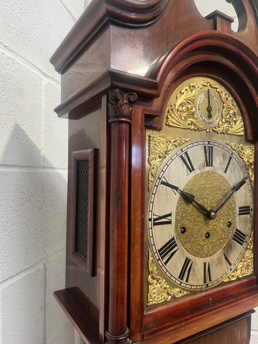 An antique Edwardian mahogany longcase clock featuring a swan-neck pediment, brass arch dial with Roman numerals, and an arched trunk door. View VI