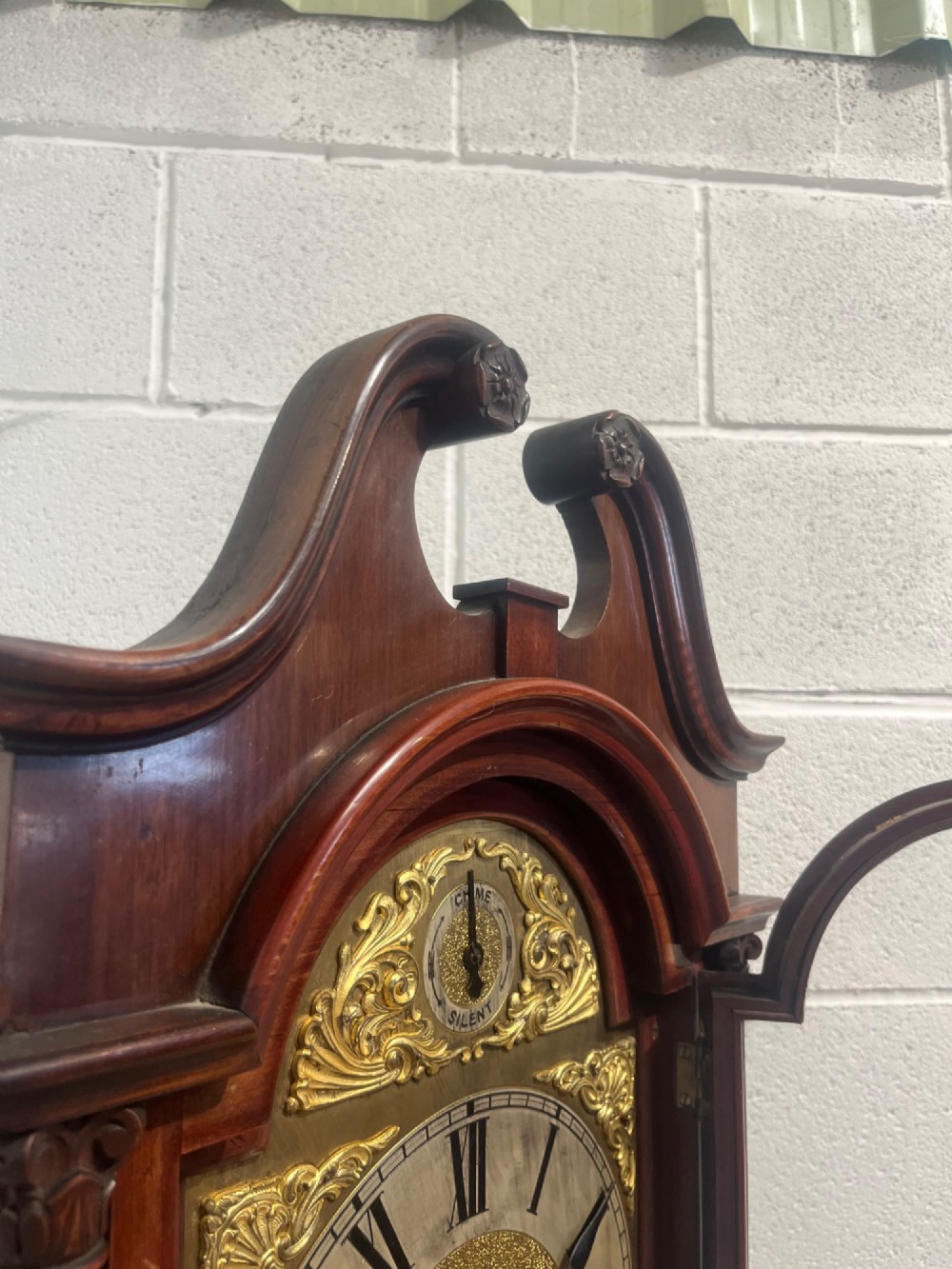 An antique Edwardian mahogany longcase clock featuring a swan-neck pediment, brass arch dial with Roman numerals, and an arched trunk door. View VIII