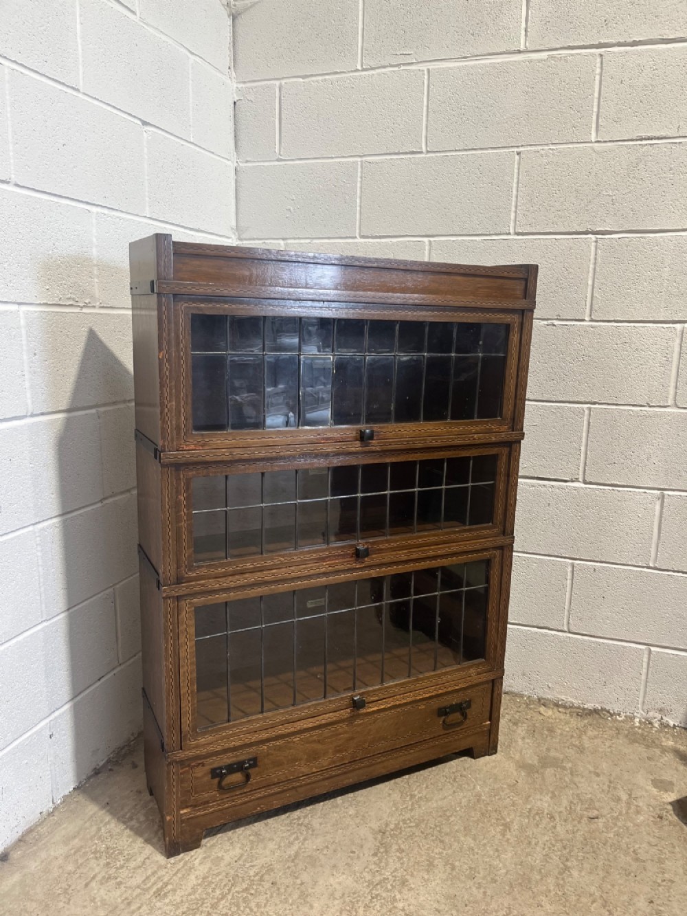 A three-tier oak sectional Globe Wernicke bookcase featuring leaded glass up-and-over doors, inlaid decorative banding, and a single bottom drawer. View V