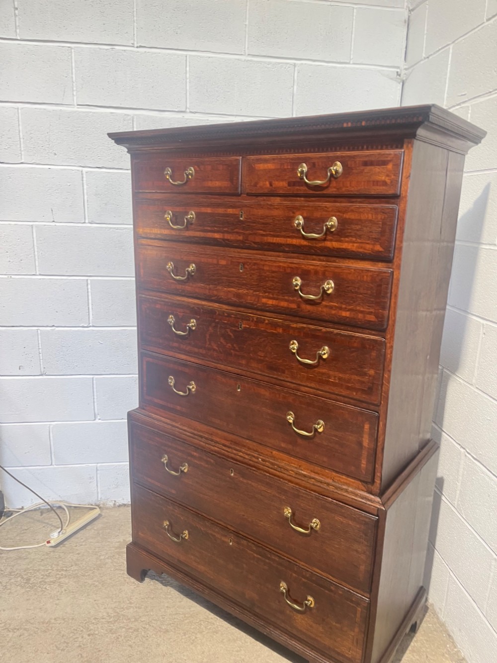 A tall George III oak chest on chest featuring brass swan-neck handles, crossbanded drawer borders, and bracket feet. View V