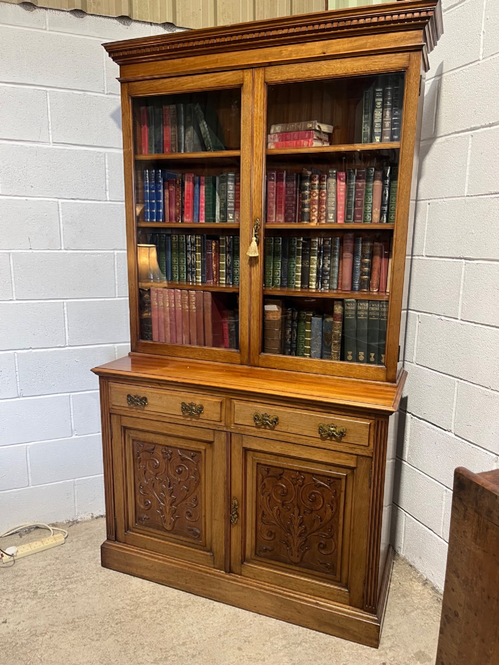 A 19th-century mahogany glazed bookcase featuring upper double glass doors filled with books, middle drawers, and lower doors with carved foliate panels. View II