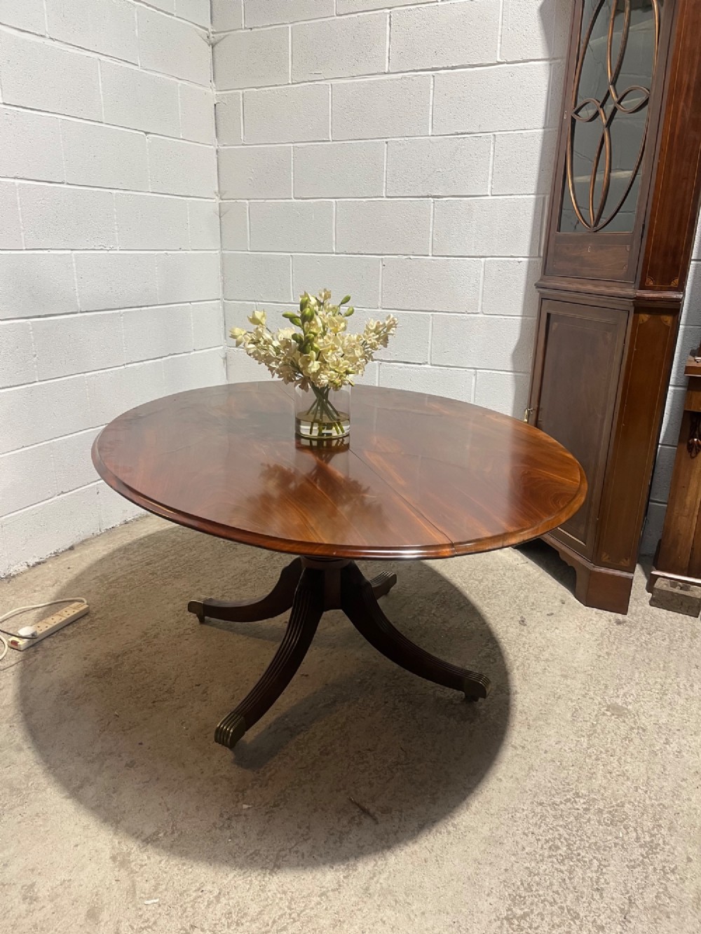A circular antique Regency mahogany dining table featuring a central turned column and four reeded splayed legs with brass castors. View II