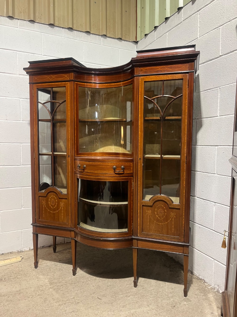 An antique inlaid mahogany display cabinet featuring a concave glazed central section, satinwood marquetry swags, astragal glazing bars, and tapered square legs.