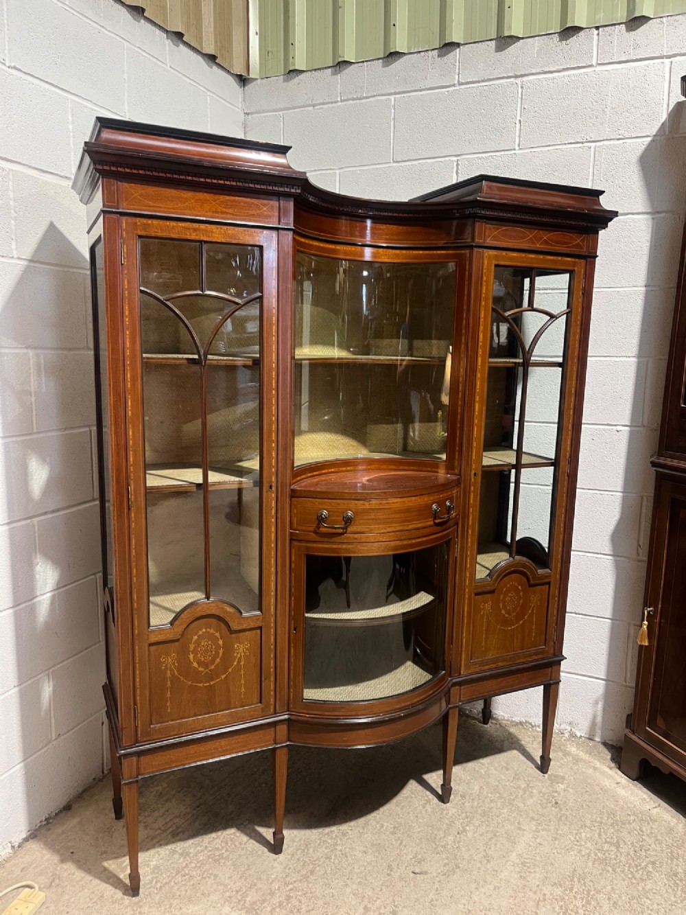 An antique inlaid mahogany display cabinet featuring a concave glazed central section, satinwood marquetry swags, astragal glazing bars, and tapered square legs. View II