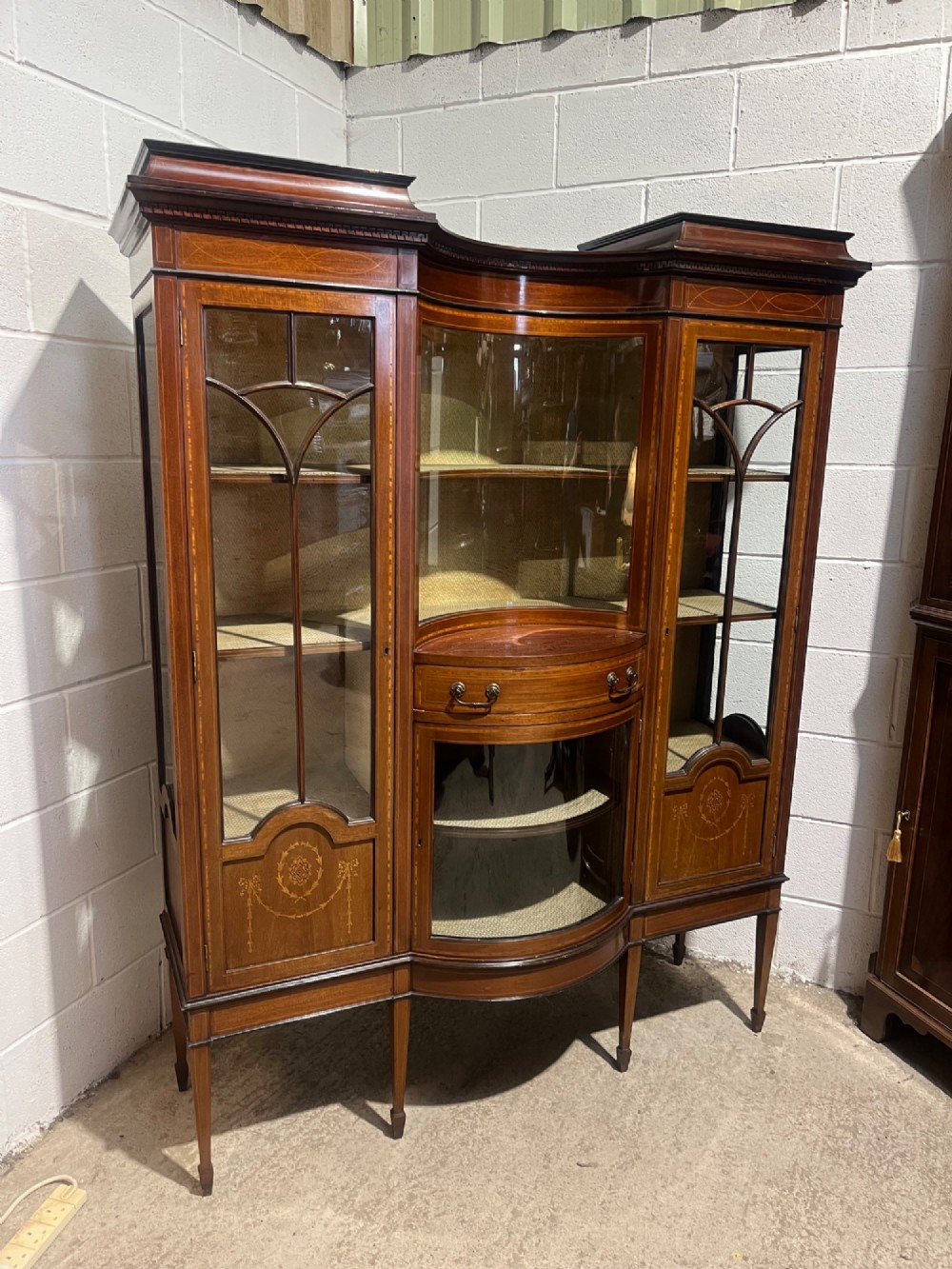 An antique inlaid mahogany display cabinet featuring a concave glazed central section, satinwood marquetry swags, astragal glazing bars, and tapered square legs. View IV