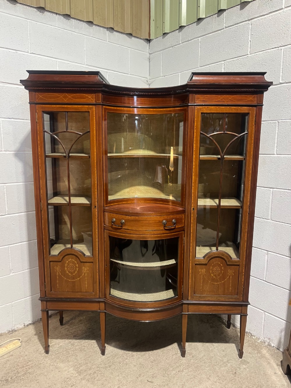 An antique inlaid mahogany display cabinet featuring a concave glazed central section, satinwood marquetry swags, astragal glazing bars, and tapered square legs. View VI