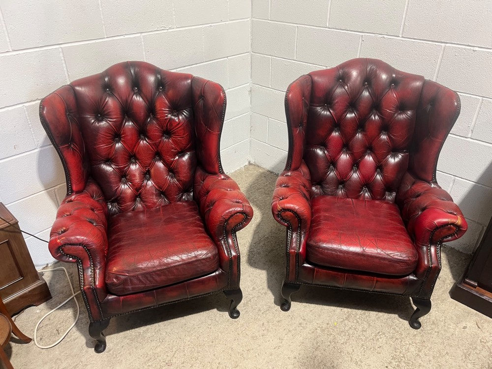 A pair of antique oxblood red leather Chesterfield wingback armchairs featuring deep button tufting, brass studding, and dark wooden cabriole front legs. View II