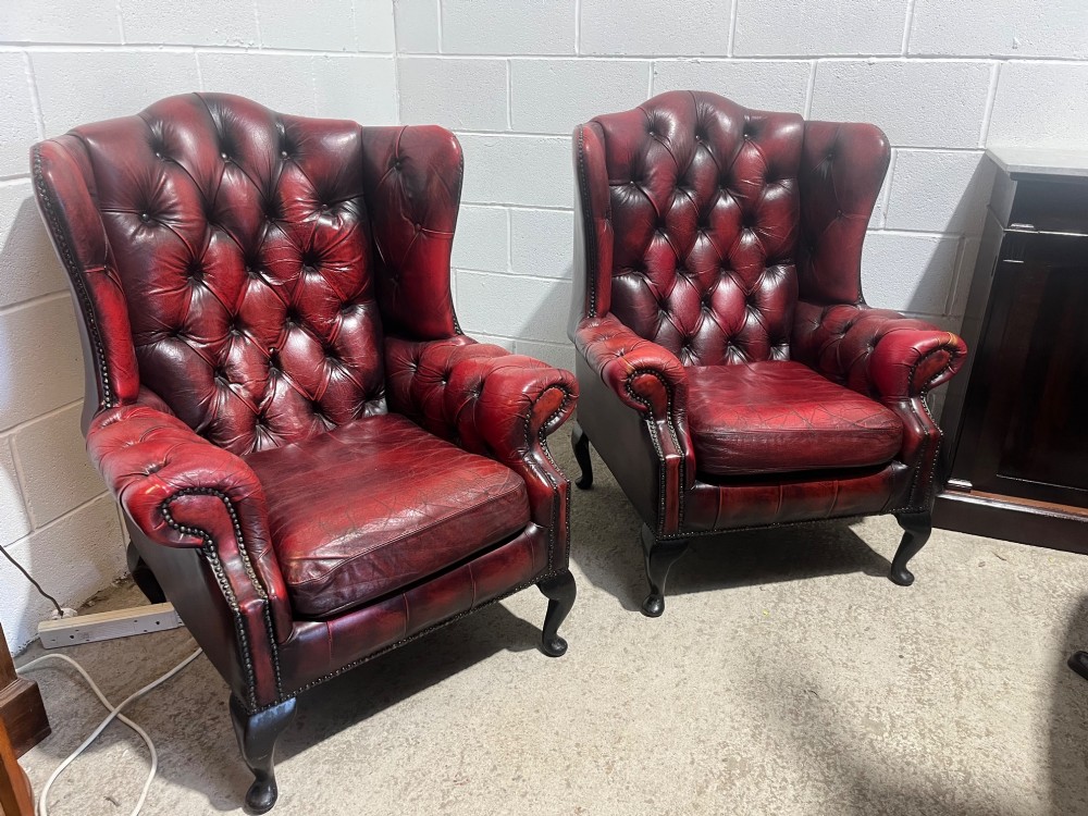 A pair of antique oxblood red leather Chesterfield wingback armchairs featuring deep button tufting, brass studding, and dark wooden cabriole front legs. View III