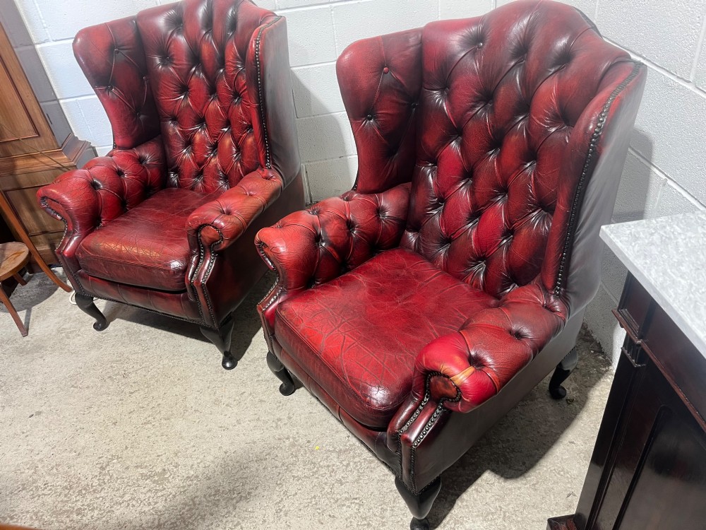 A pair of antique oxblood red leather Chesterfield wingback armchairs featuring deep button tufting, brass studding, and dark wooden cabriole front legs. View V
