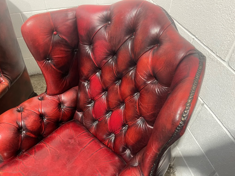 A pair of antique oxblood red leather Chesterfield wingback armchairs featuring deep button tufting, brass studding, and dark wooden cabriole front legs. View VI