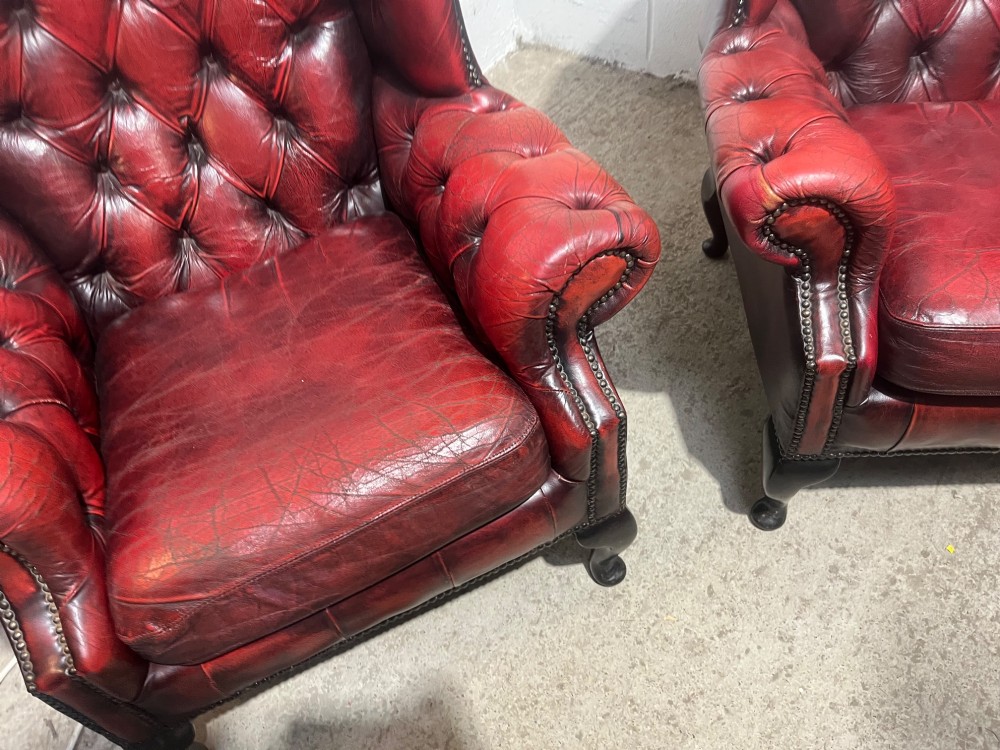 A pair of antique oxblood red leather Chesterfield wingback armchairs featuring deep button tufting, brass studding, and dark wooden cabriole front legs. View VII