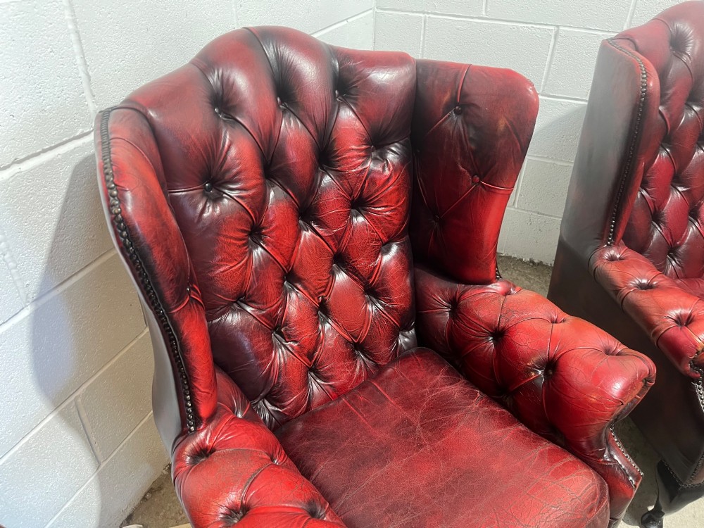 A pair of antique oxblood red leather Chesterfield wingback armchairs featuring deep button tufting, brass studding, and dark wooden cabriole front legs. View X