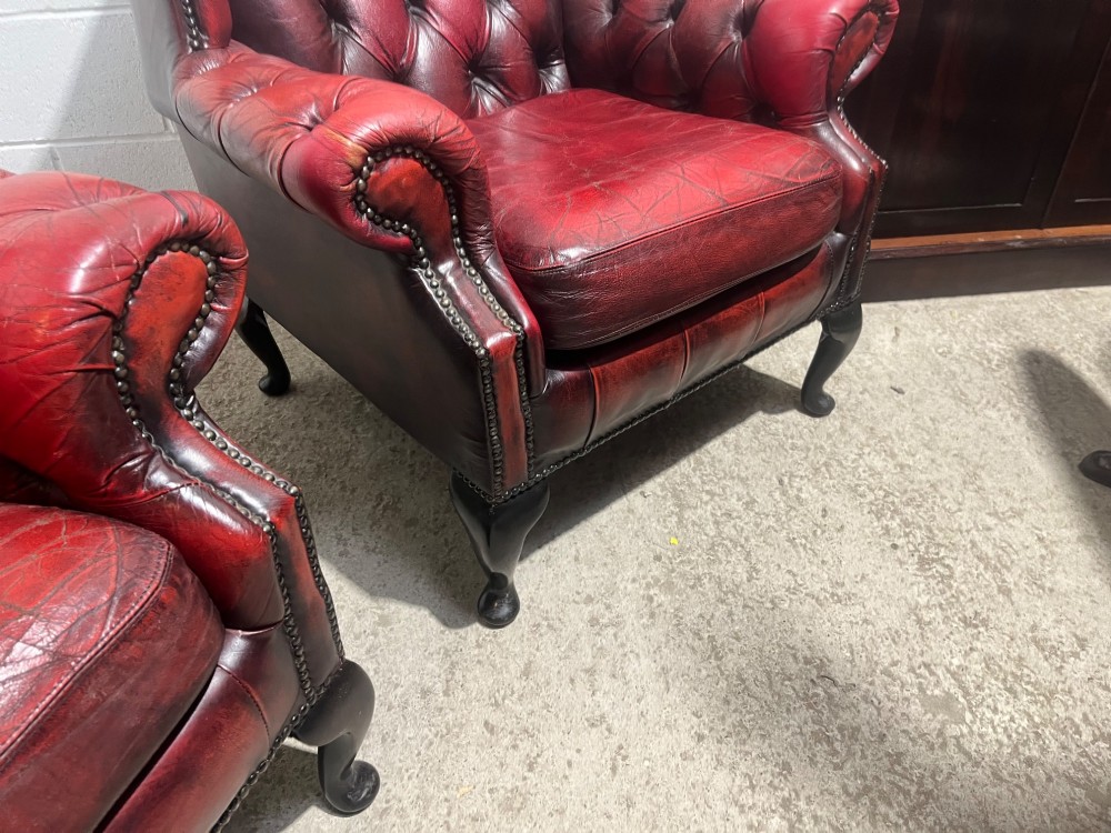 A pair of antique oxblood red leather Chesterfield wingback armchairs featuring deep button tufting, brass studding, and dark wooden cabriole front legs. View XI