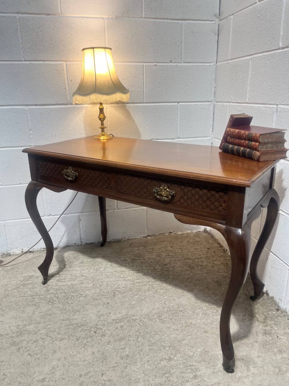 An Edwardian dark mahogany writing desk featuring two blind fretwork carved drawers with brass drop handles and slender cabriole legs on brass castors. View VI
