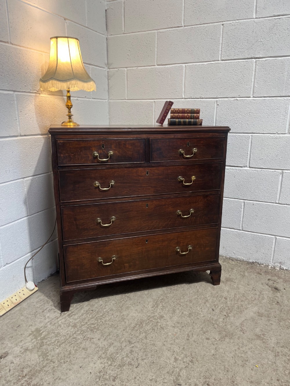 An antique Georgian dark oak chest of drawers with two short drawers above three long drawers, featuring brass swan-neck handles and bracket feet. View II