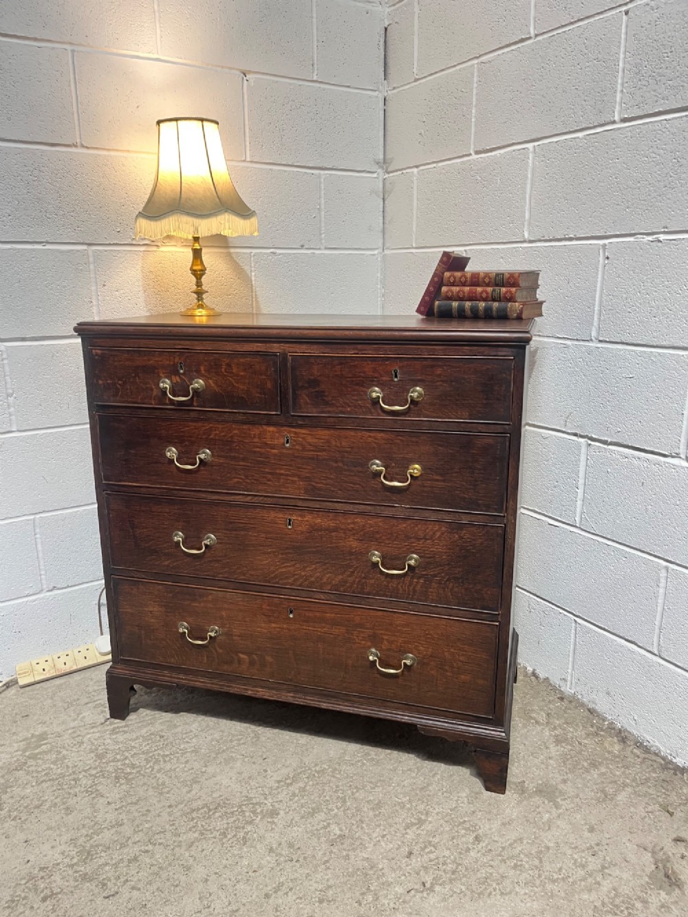 An antique Georgian dark oak chest of drawers with two short drawers above three long drawers, featuring brass swan-neck handles and bracket feet. View III