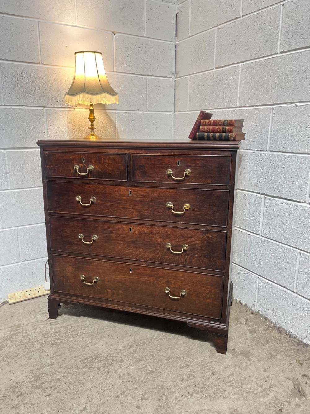 An antique Georgian dark oak chest of drawers with two short drawers above three long drawers, featuring brass swan-neck handles and bracket feet. View IV