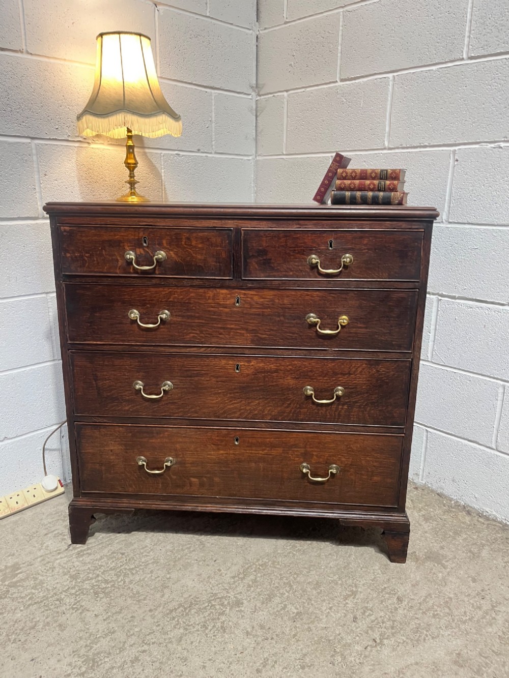 An antique Georgian dark oak chest of drawers with two short drawers above three long drawers, featuring brass swan-neck handles and bracket feet. View V