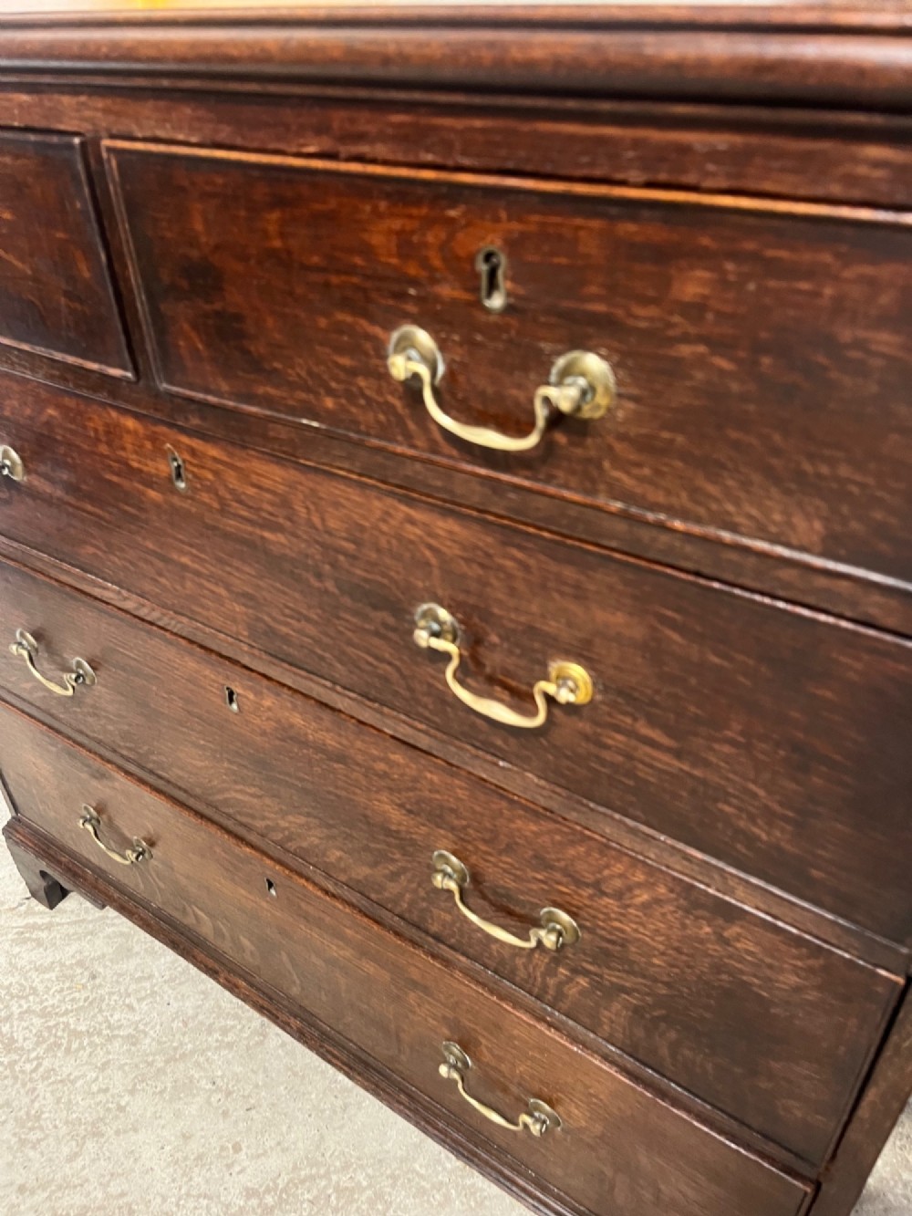 An antique Georgian dark oak chest of drawers with two short drawers above three long drawers, featuring brass swan-neck handles and bracket feet. View VII