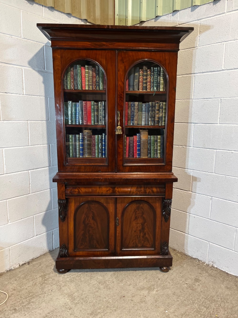 A 19th-century mahogany bookcase featuring two upper arched glazed doors displaying books, resting upon a lower two-door cupboard with flame veneer panels and carved scroll brackets. View II