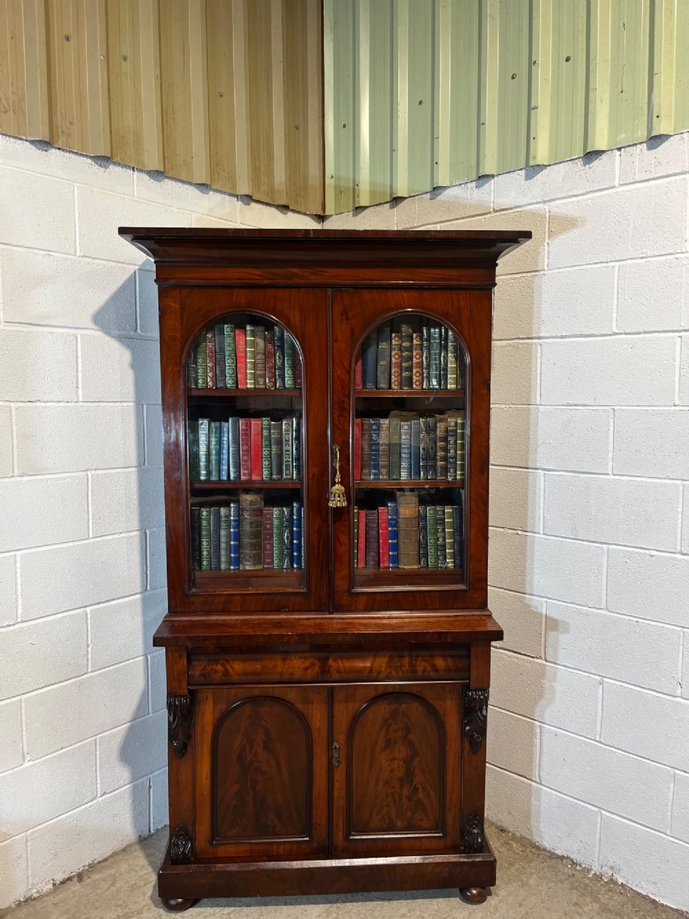 A 19th-century mahogany bookcase featuring two upper arched glazed doors displaying books, resting upon a lower two-door cupboard with flame veneer panels and carved scroll brackets. View V