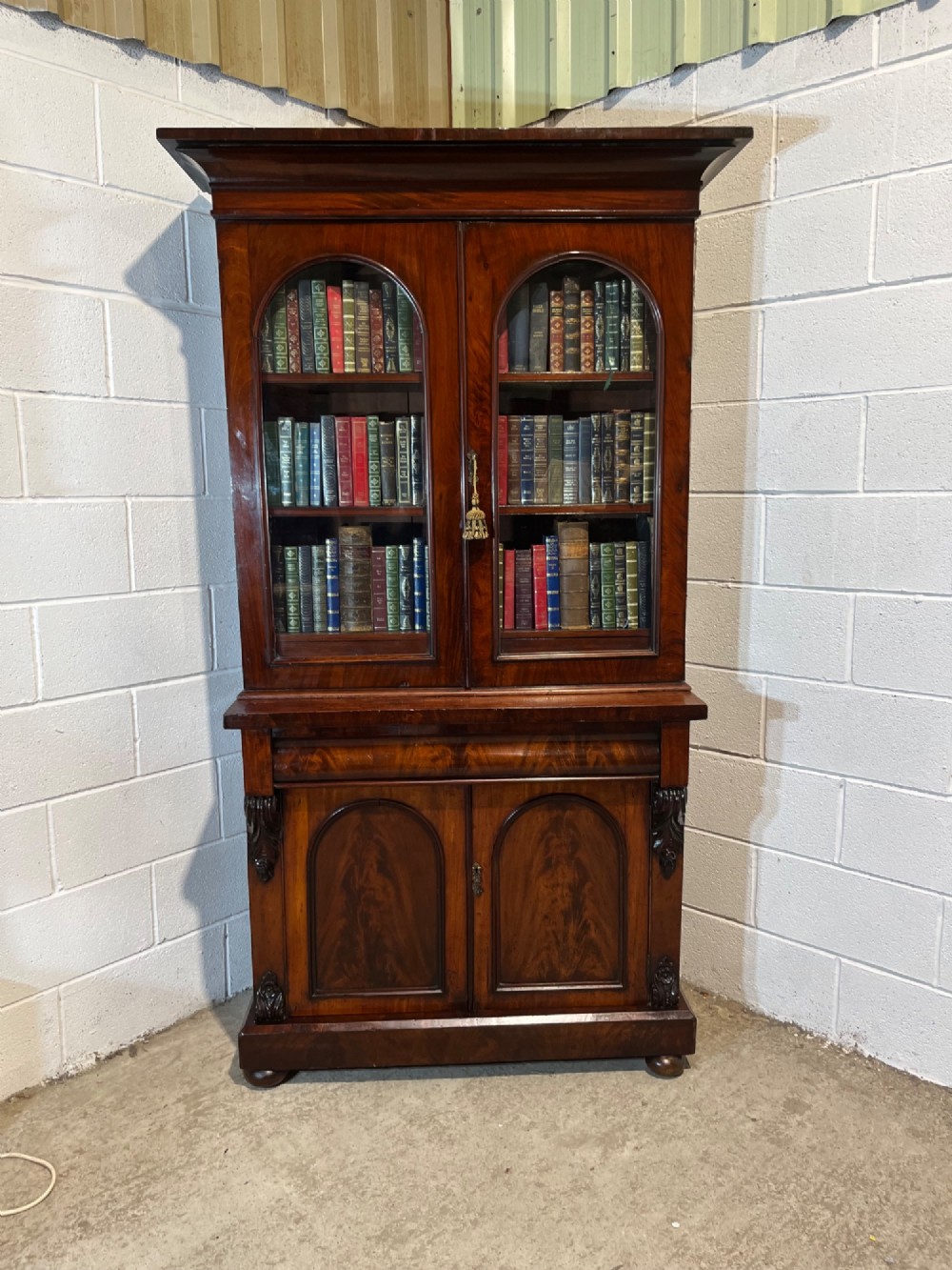 A 19th-century mahogany bookcase featuring two upper arched glazed doors displaying books, resting upon a lower two-door cupboard with flame veneer panels and carved scroll brackets. View VI