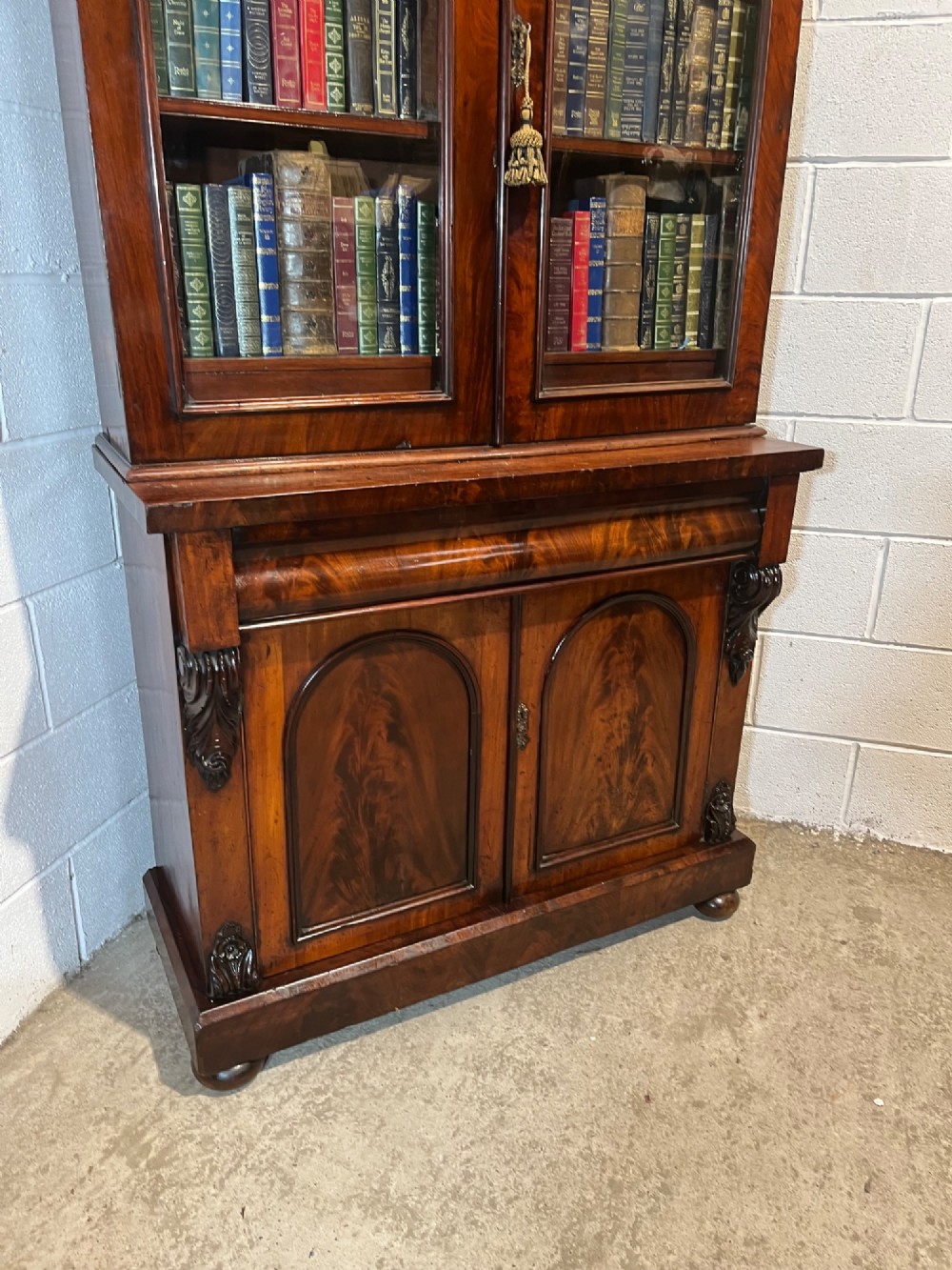 A 19th-century mahogany bookcase featuring two upper arched glazed doors displaying books, resting upon a lower two-door cupboard with flame veneer panels and carved scroll brackets. View VII