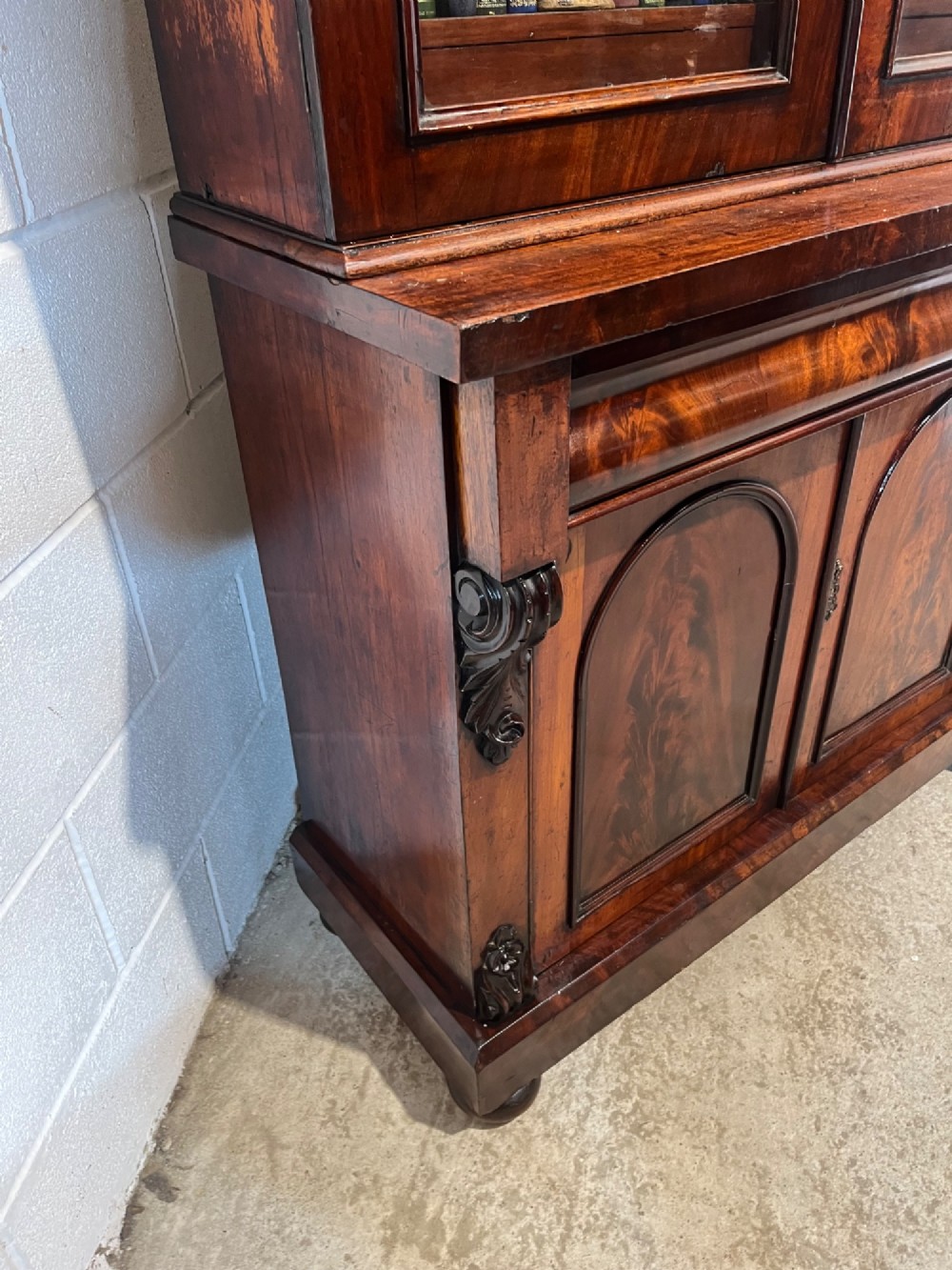 A 19th-century mahogany bookcase featuring two upper arched glazed doors displaying books, resting upon a lower two-door cupboard with flame veneer panels and carved scroll brackets. View VIII