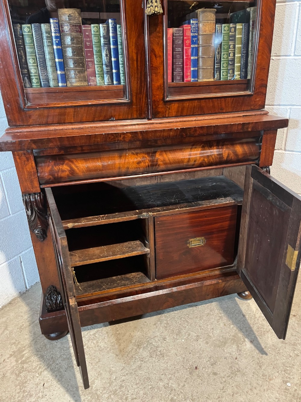 A 19th-century mahogany bookcase featuring two upper arched glazed doors displaying books, resting upon a lower two-door cupboard with flame veneer panels and carved scroll brackets. View XI