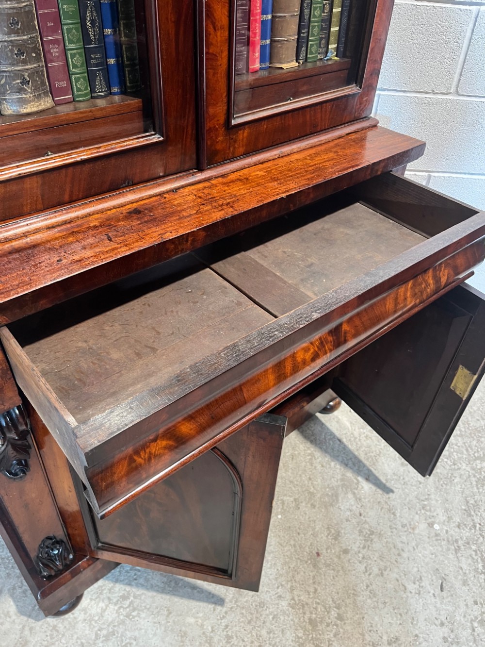 A 19th-century mahogany bookcase featuring two upper arched glazed doors displaying books, resting upon a lower two-door cupboard with flame veneer panels and carved scroll brackets. View XII