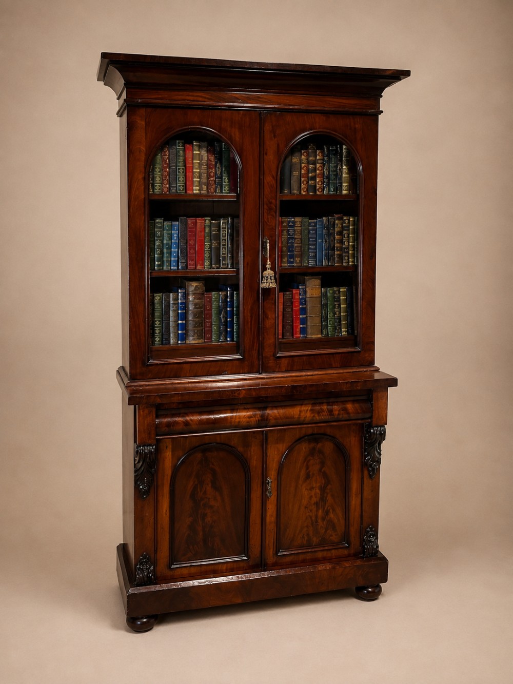 A 19th-century mahogany bookcase featuring two upper arched glazed doors displaying books, resting upon a lower two-door cupboard with flame veneer panels and carved scroll brackets.