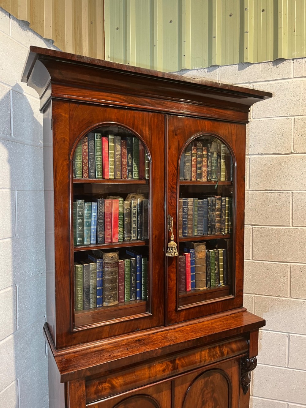 A 19th-century mahogany bookcase featuring two upper arched glazed doors displaying books, resting upon a lower two-door cupboard with flame veneer panels and carved scroll brackets. View VII