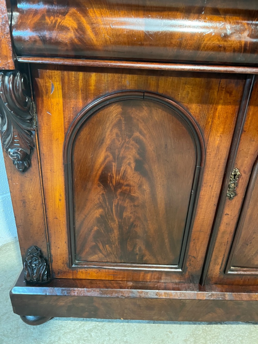 A 19th-century mahogany bookcase featuring two upper arched glazed doors displaying books, resting upon a lower two-door cupboard with flame veneer panels and carved scroll brackets. View XIII