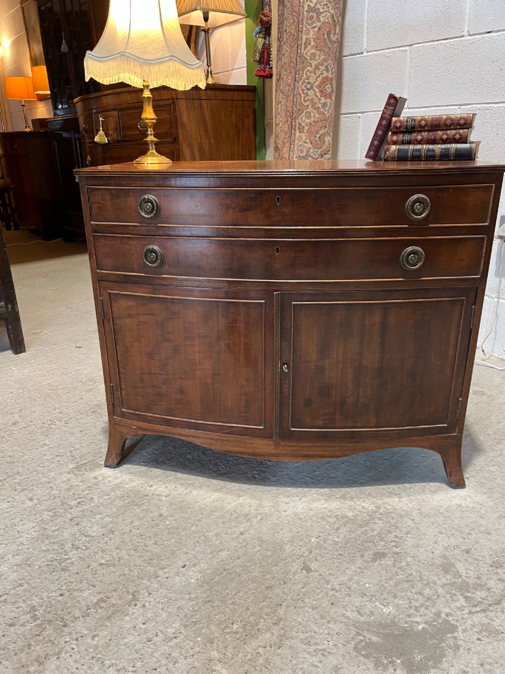 An early 20th-century mahogany bowfront cupboard with two top drawers featuring brass ring handles above a double-door lower compartment. View V