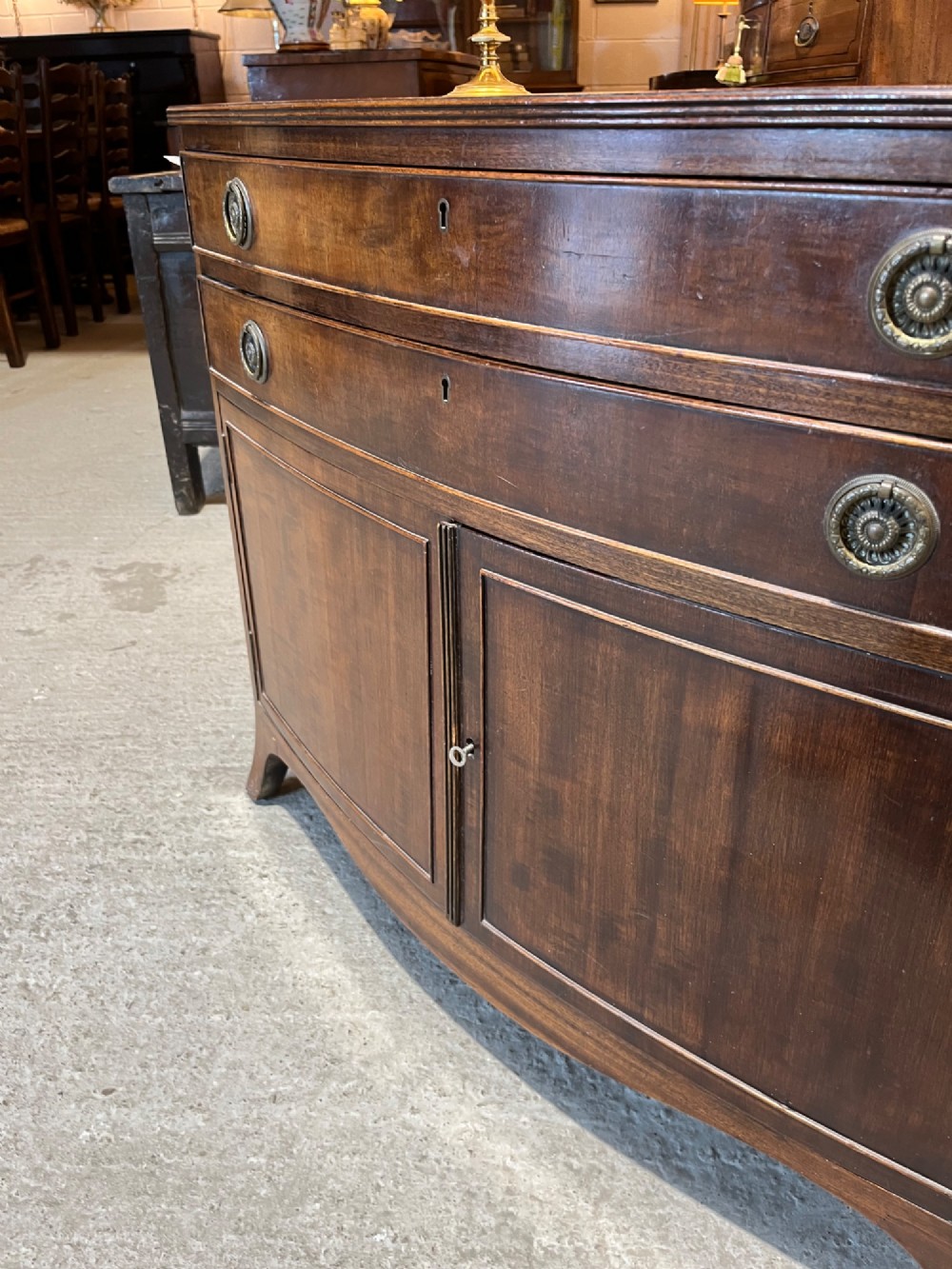 An early 20th-century mahogany bowfront cupboard with two top drawers featuring brass ring handles above a double-door lower compartment. View VI