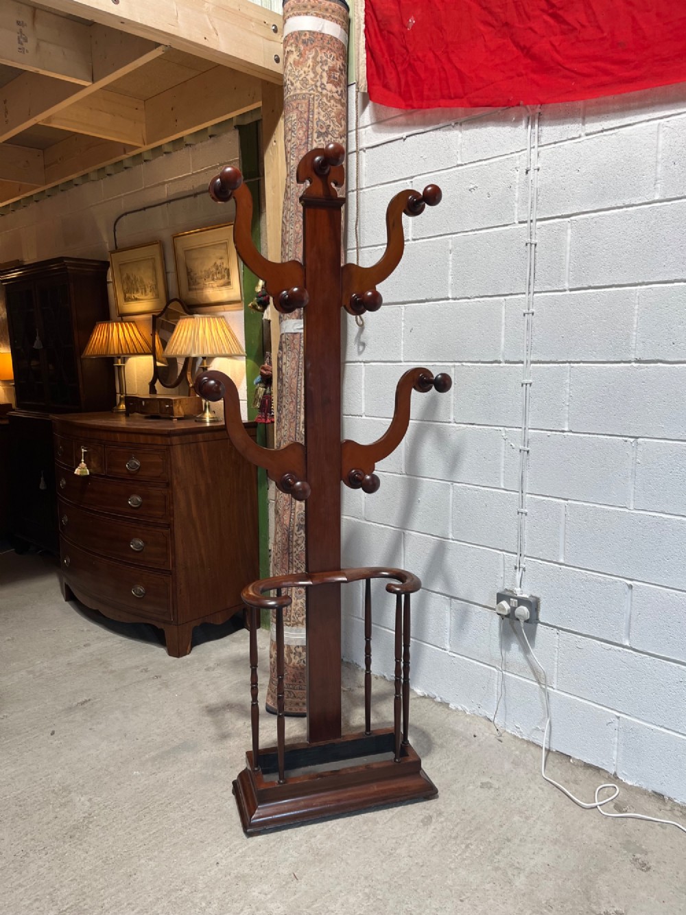 A 19th-century mahogany freestanding hall stand featuring turned wooden pegs on curved scroll arms above a lower umbrella holder with a drip tray. View X
