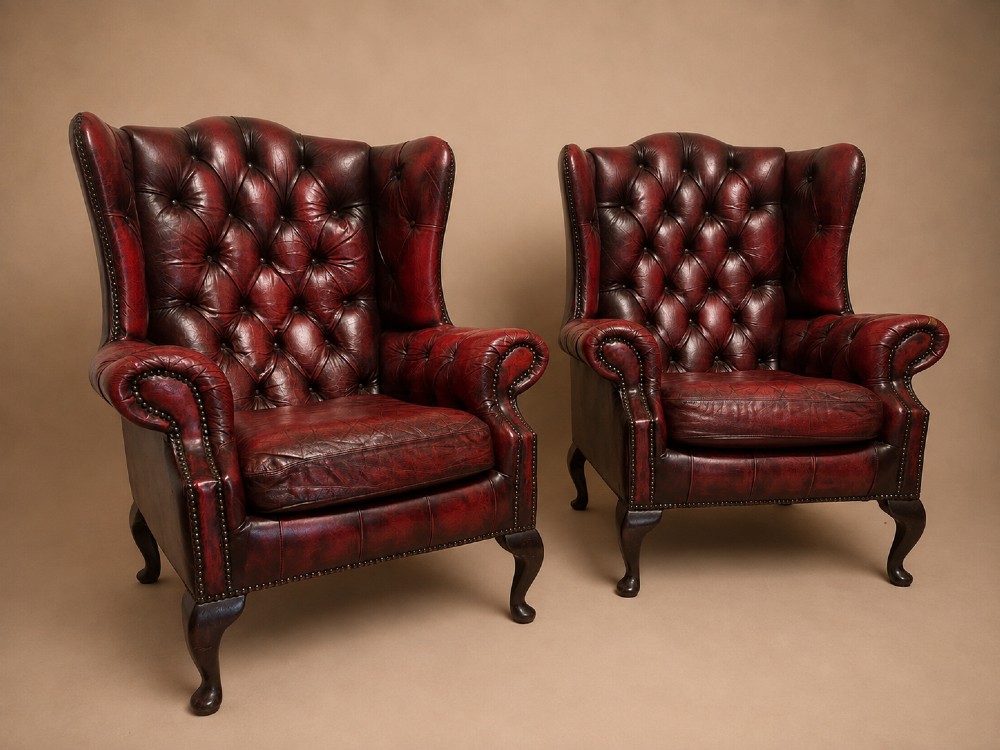 A pair of antique oxblood red leather Chesterfield wingback armchairs featuring deep button tufting, brass studding, and dark wooden cabriole front legs.