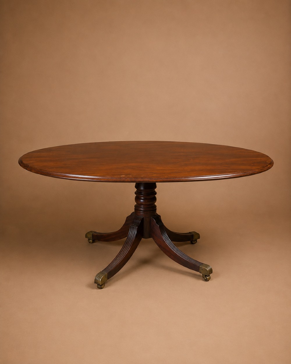 An oval Regency mahogany dining table featuring a central turned pedestal supported by four reeded sabre legs with brass casters.