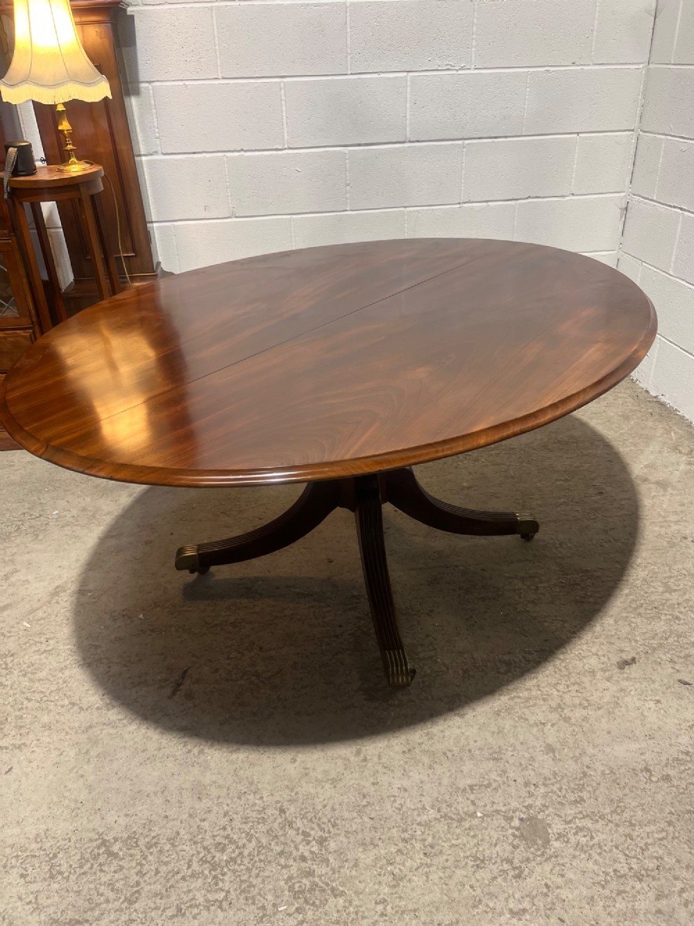 An oval Regency mahogany dining table featuring a central turned pedestal supported by four reeded sabre legs with brass casters. View IX