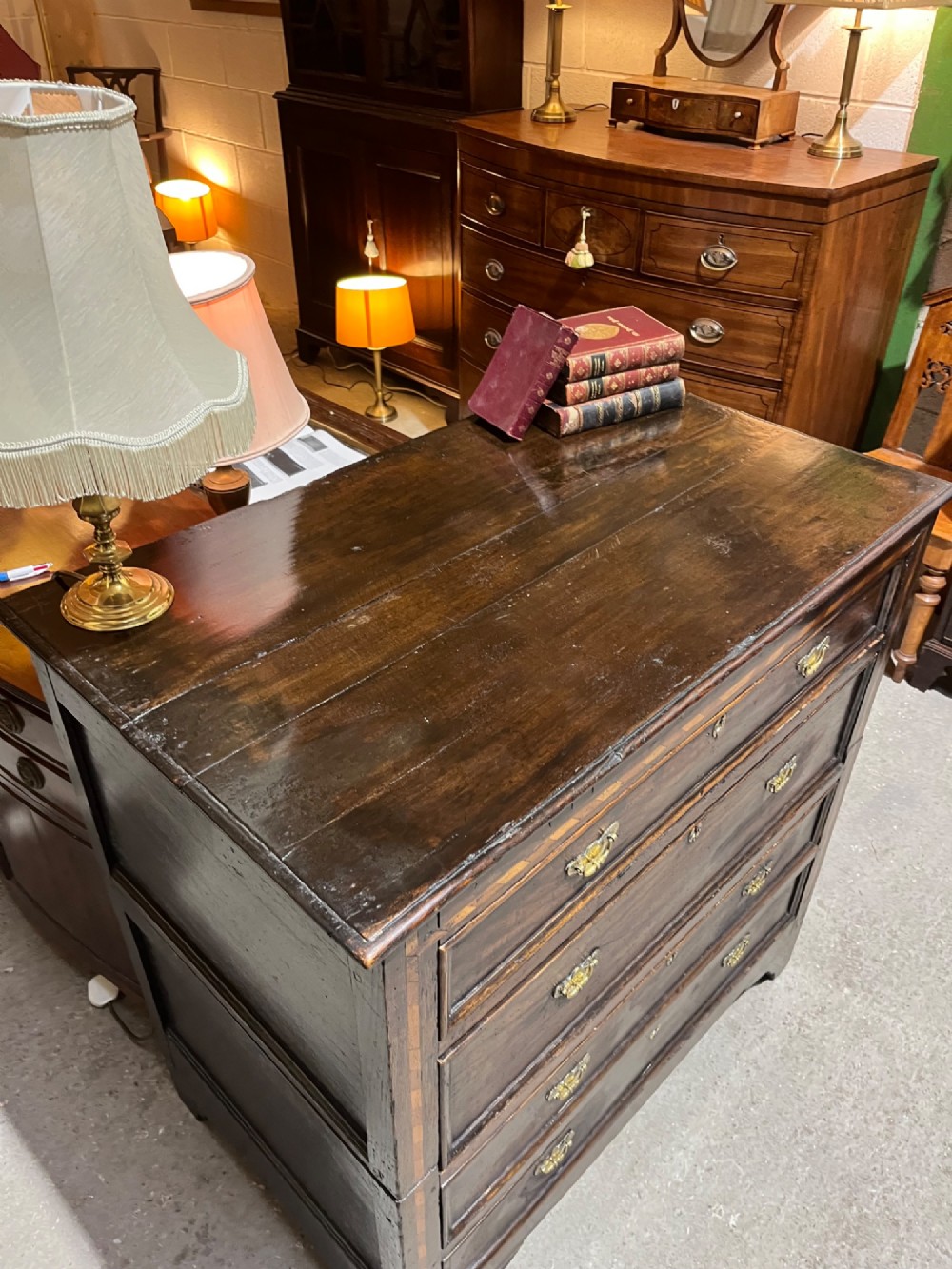 An early 18th-century dark oak chest of drawers featuring geometric inlay along the outer frame, four graduated drawers with brass handles, and bracket feet. View VI