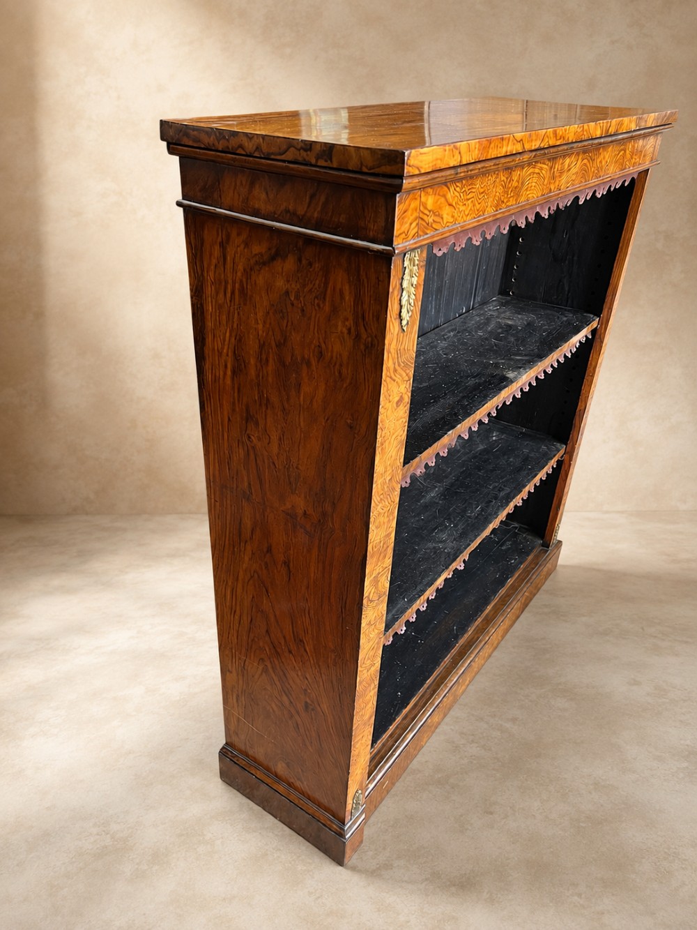 A 19th-century open walnut bookcase featuring highly figured veneer, brass corner mounts, three shelves with scalloped trim, and a dark painted back panel. View VI