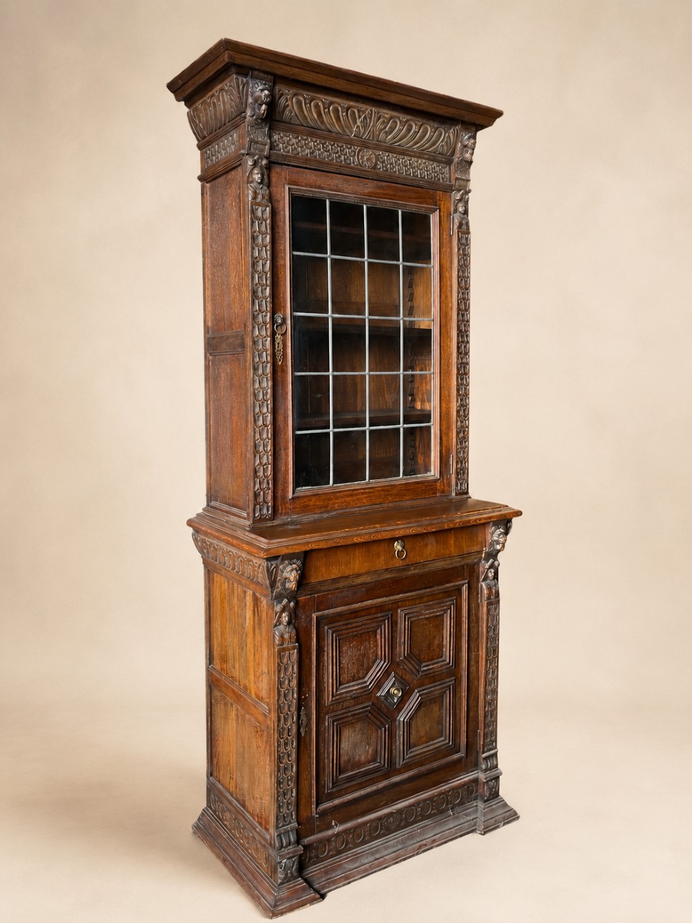 A slim 19th-century carved oak bookcase featuring a single leaded glass upper door, a central drawer, and a lower carved cupboard door with geometric panels. View II