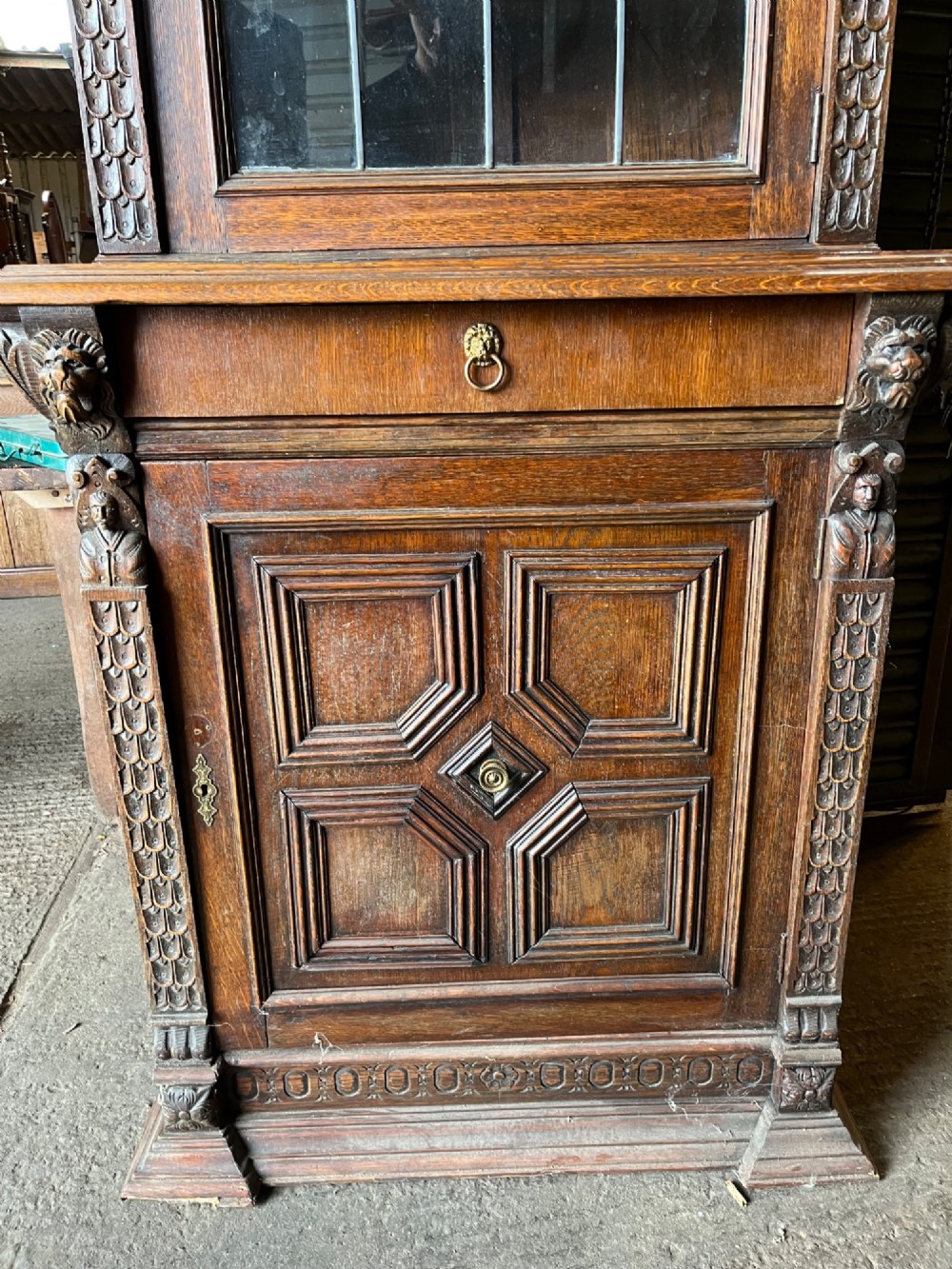 A slim 19th-century carved oak bookcase featuring a single leaded glass upper door, a central drawer, and a lower carved cupboard door with geometric panels. View IV