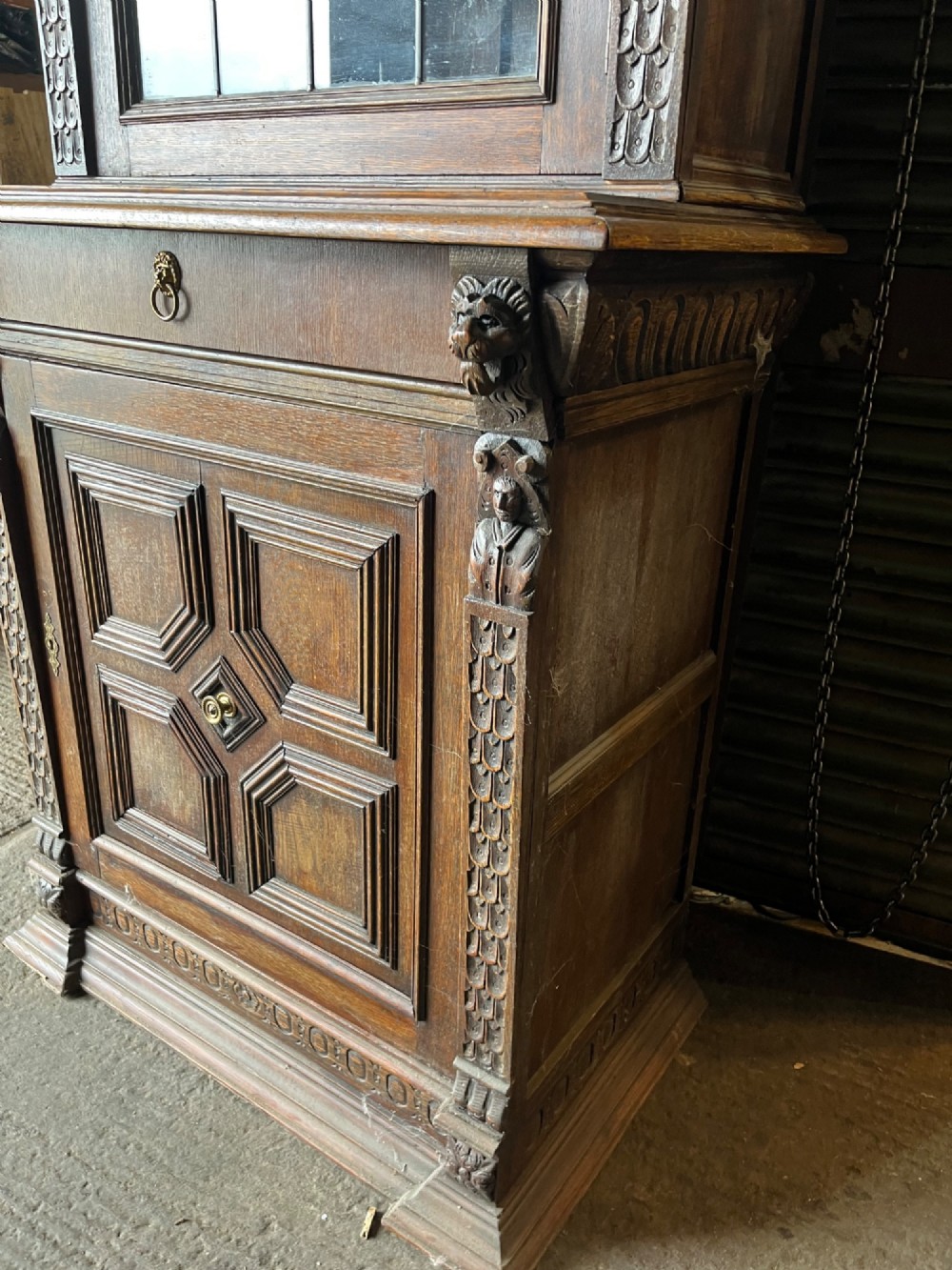 A slim 19th-century carved oak bookcase featuring a single leaded glass upper door, a central drawer, and a lower carved cupboard door with geometric panels. View V